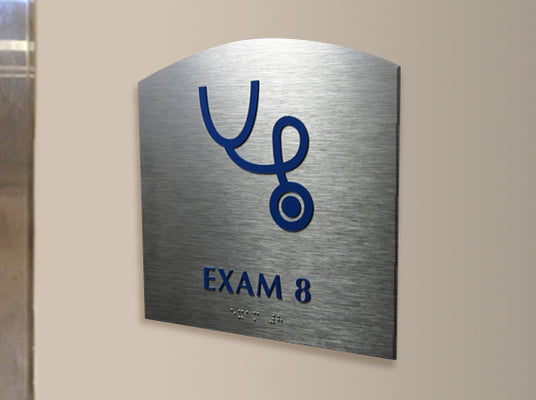 Metal wall identification sign with a stethoscope symbol and 'EXAM 8' text and Braille on a beige wall.