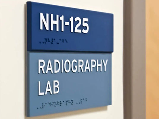 Blue sign with 'NH1-125 Radiography Lab' tactile text and Braille on a wall.
