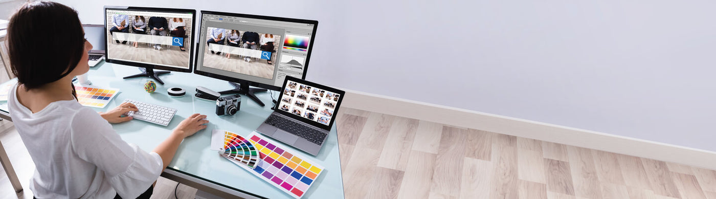 Graphic designer working at a desk with multiple computer monitors, a laptop, and color books.