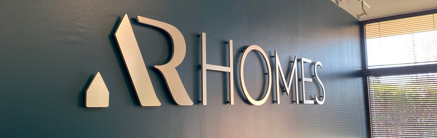 Dimensional metallic letters and logo of AR Homes on a wall with a window in the background