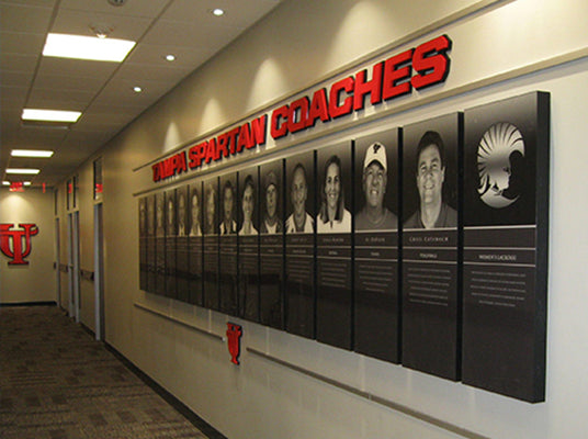 Wall with 'YSU Spartan Coaches' acrylic recognition plaques and portraits of coaches in a hallway.