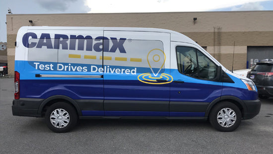 Van with Carmax branding on a full vehicle wrap, parked in a parking lot.