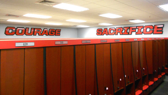 Row of locker doors with acrylic dimensional letters reading 'COURAGE' and 'SACRIFICE' above them in a sports facility.
