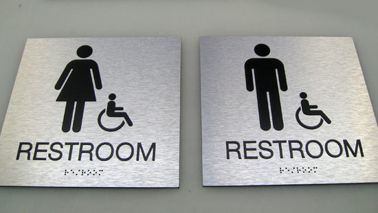 Two metallic ADA restroom signs with accessibility symbols and tactile letters and Braille text reading "Restrom,' on a gray background