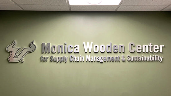 Interior dimensional metal sign with 'Monica Wooden Center for Supply Chain Management & Sustainability' on a green wall.