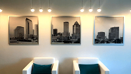 Three framed cityscape posters on a wall above two white chairs with green pillows.