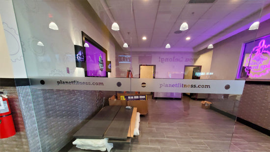 Interior of a Planet Fitness gym with workout equipment and digital screens, and branded window film applied to a glass wall.