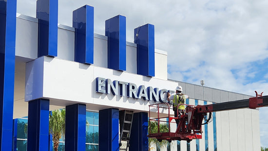 Sign-Age employee on a cherry picker installing an 'Entrance' sign on a building with blue and white design.