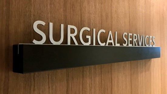 Surgical Services Aluminum letters sign on a shelf support mounted on a paneled wall.