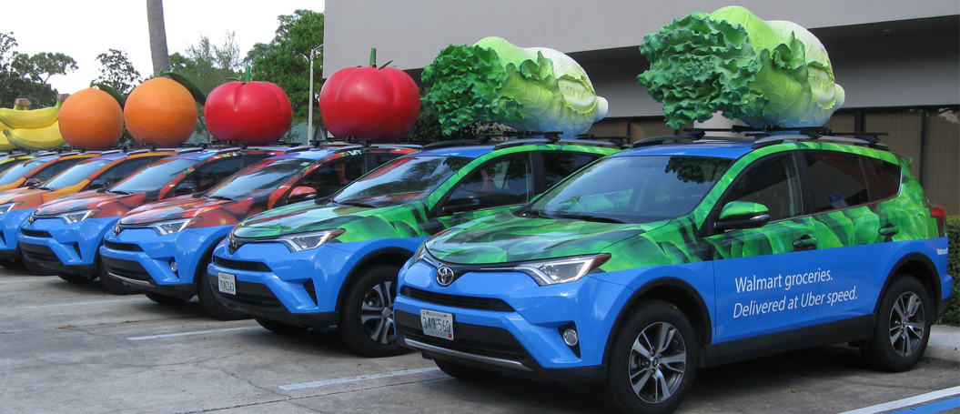 Row of colorful delivery vehicles with oversized produce models on top, parked in a lot.
