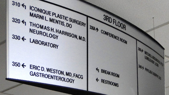 Overhead directional sign in a medical facility with various room and department names.