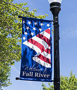American flag pole banner with 'Welcome to Fall River' text on a pole against a blue sky.